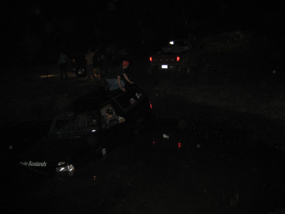 Dark SUV stuck in a pond at night with men on it and a Ford pickup on the bank