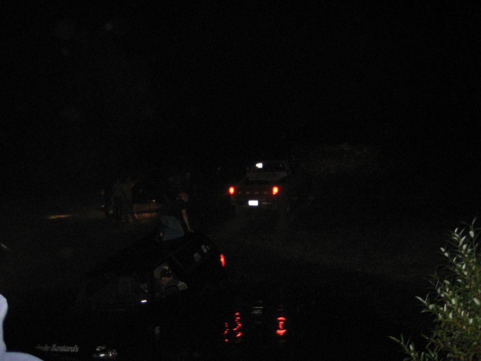 Night photo of a vehicle submerged in a pond with a truck and onlookers on the bank