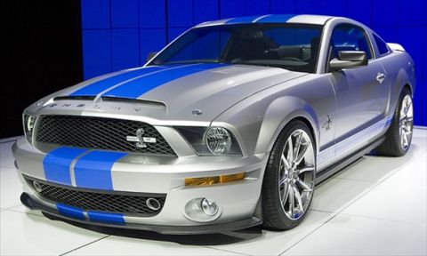1996 Ford Bronco - A silver Ford Mustang Shelby GT500 with blue racing stripes is displayed on a white platform with a blue background.