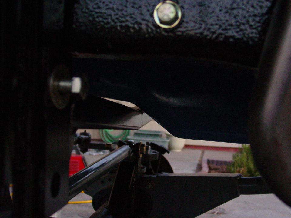 Close-up of dark vehicle suspension parts, including a metal bar and bolts, with a water hose visible in the background