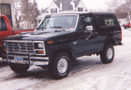 Black 1986 Ford Bronco parked in snow next to a red truck