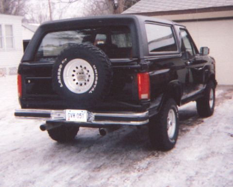 1986 Ford Bronco - This black Ford Bronco is parked outside a garage in a snowy environment, showing its rear profile and spare tire.
