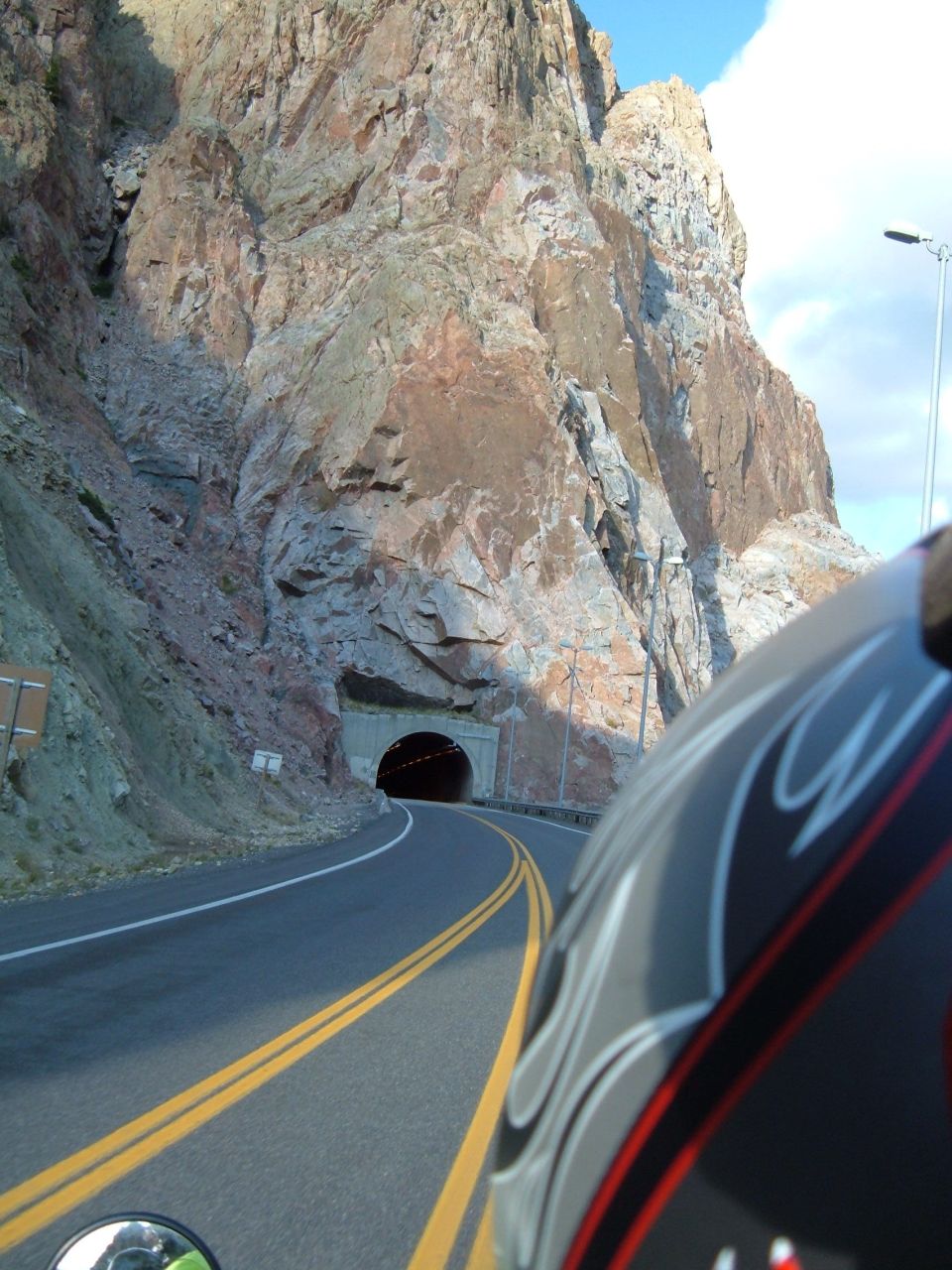 Riding a motorcycle towards a tunnel entrance in a rocky mountain pass. Part of the helmet is visible