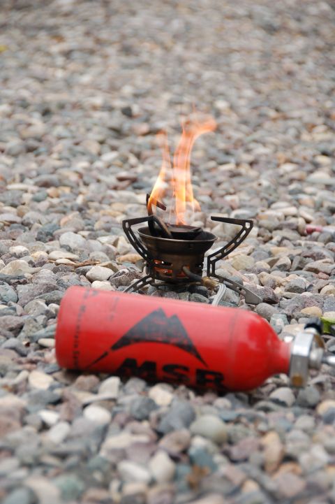 1998 BMW R1100GS ABS - A red MSR fuel canister lies next to a camping stove with flames on a bed of gravel.