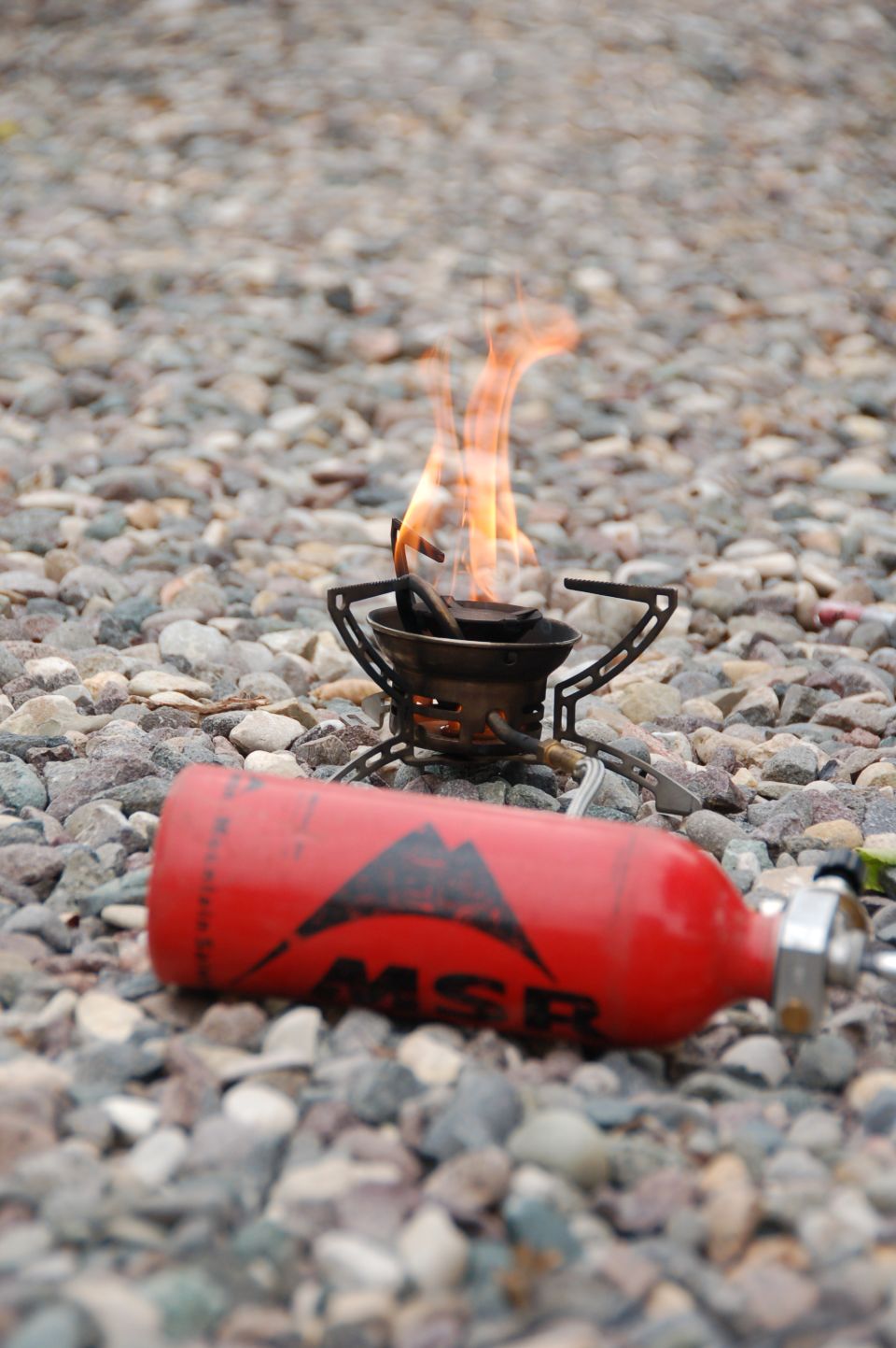 Red MSR fuel canister and camping stove with flames on gravel