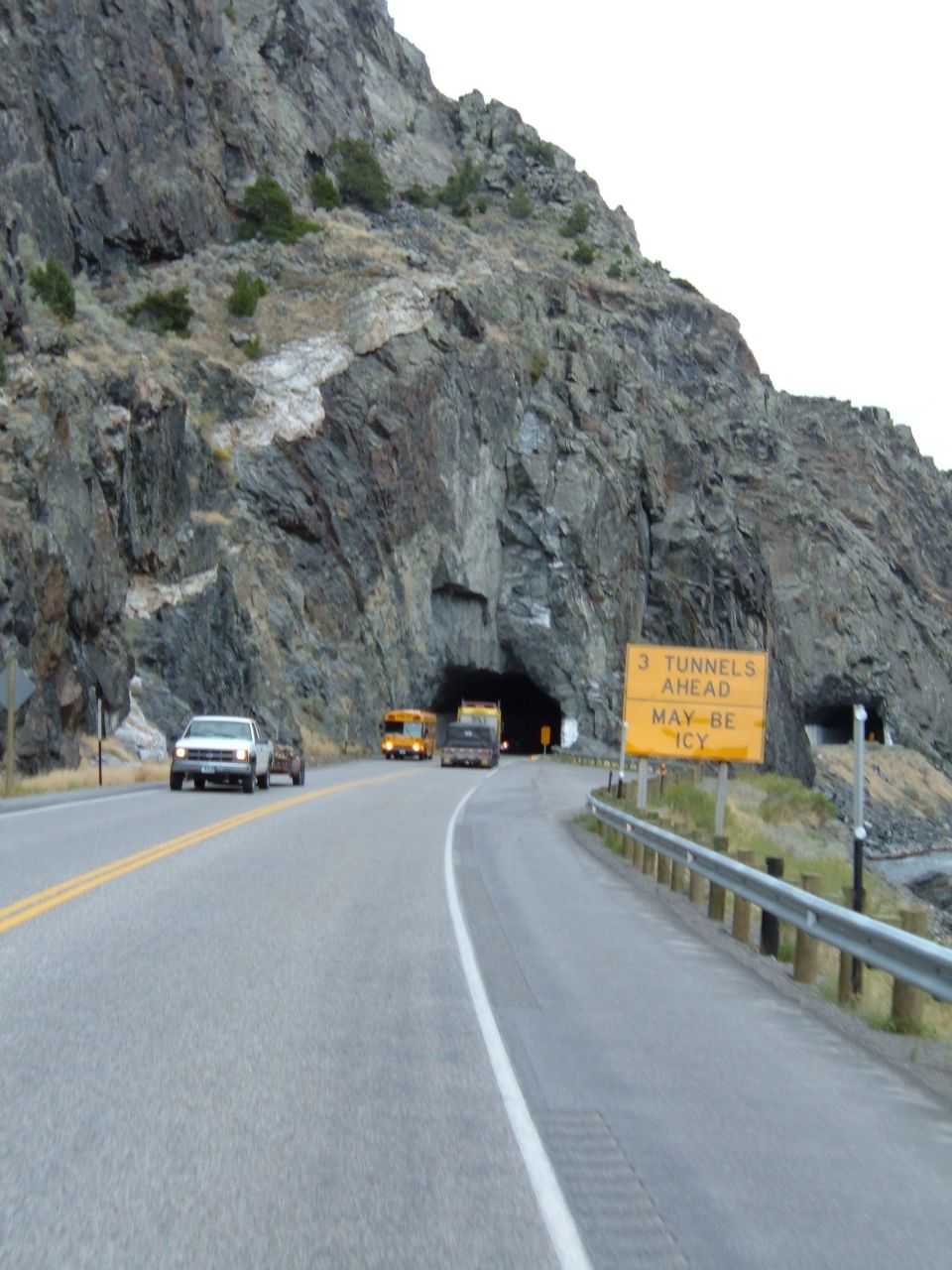 Road leading to a tunnel entrance in a rocky mountainside, with a white pickup truck, a yellow school bus, and other vehicles visible
