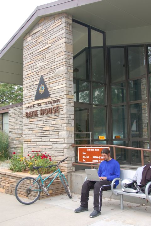 1998 BMW R1100GS ABS - A person sits on a bench outside the State Bath House, using a laptop, with a bicycle parked next to some flowers.