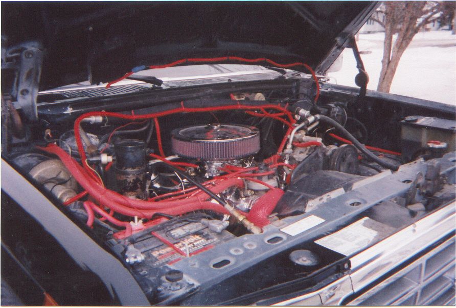 Engine bay of dark Ford Bronco with chrome air cleaner, red plug wires and hoses, PowerVolt battery
