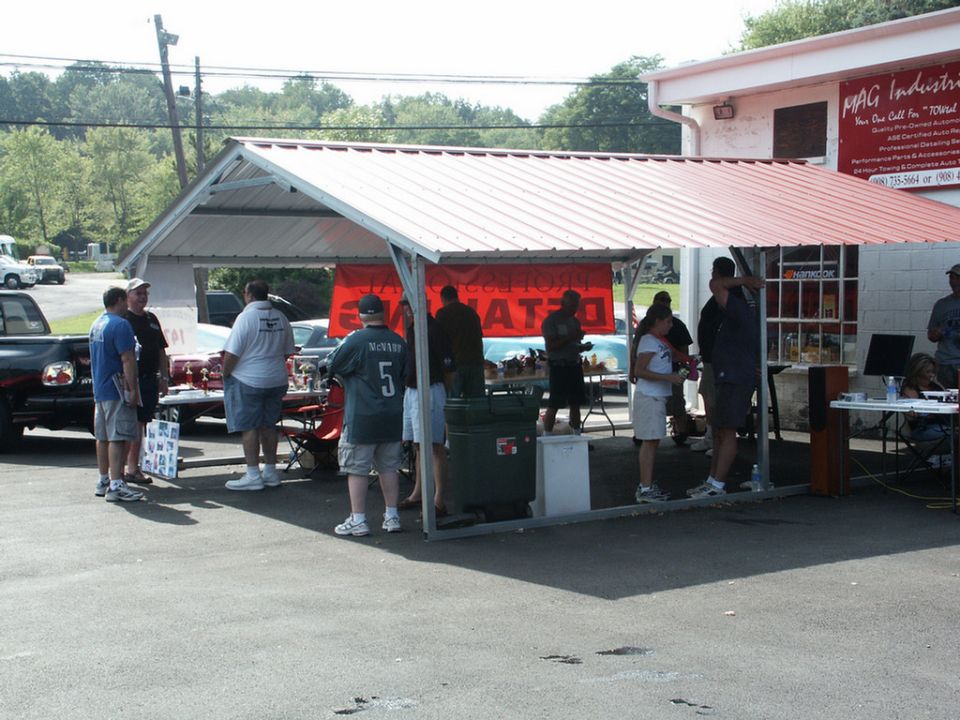 Outdoor car show with tents, tables, and people gathered. A red banner hangs in the background
