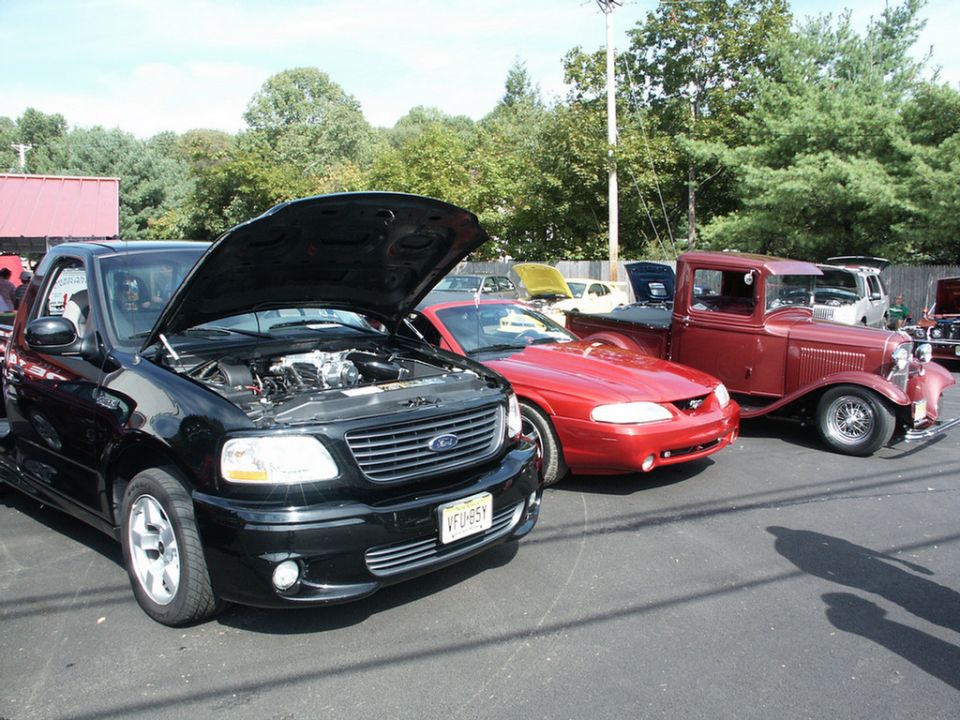 Black 2001 Ford F-150 Lightning pickup truck, hood open showing engine. Red Ford Mustang and maroon 1930s pickup truck also visible