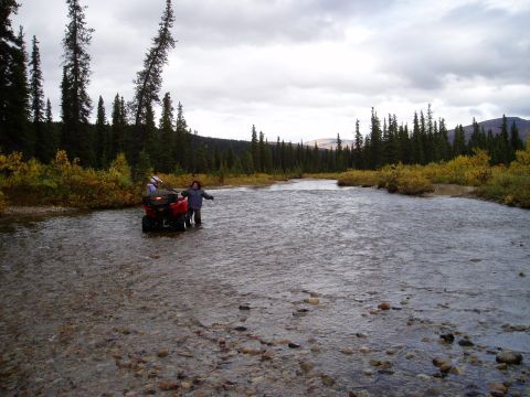 2000 Ford E350 - Two individuals navigate a red ATV through a rocky riverbed surrounded by dense forest and autumn foliage.