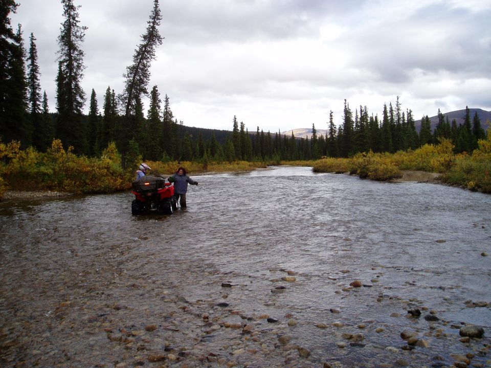 Red ATV with two people crossing a river in a wooded area