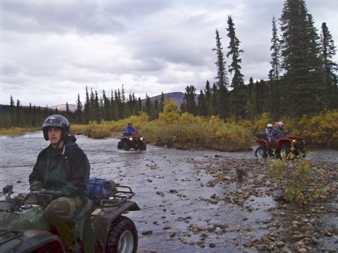 2000 Ford E350 - ATV riders navigate a river in a scenic, wooded landscape under a cloudy sky.
