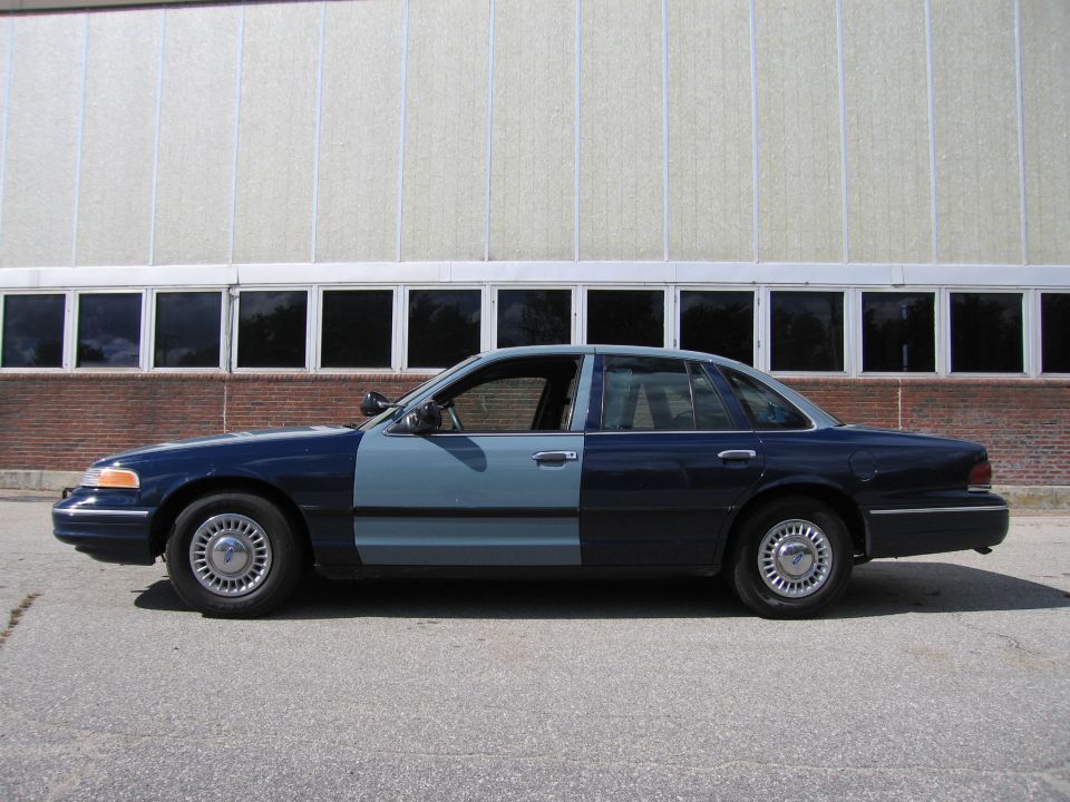 Side view of a two-tone Ford Crown Victoria, dark blue rear half and light blue front half. Parked on asphalt in front of a building