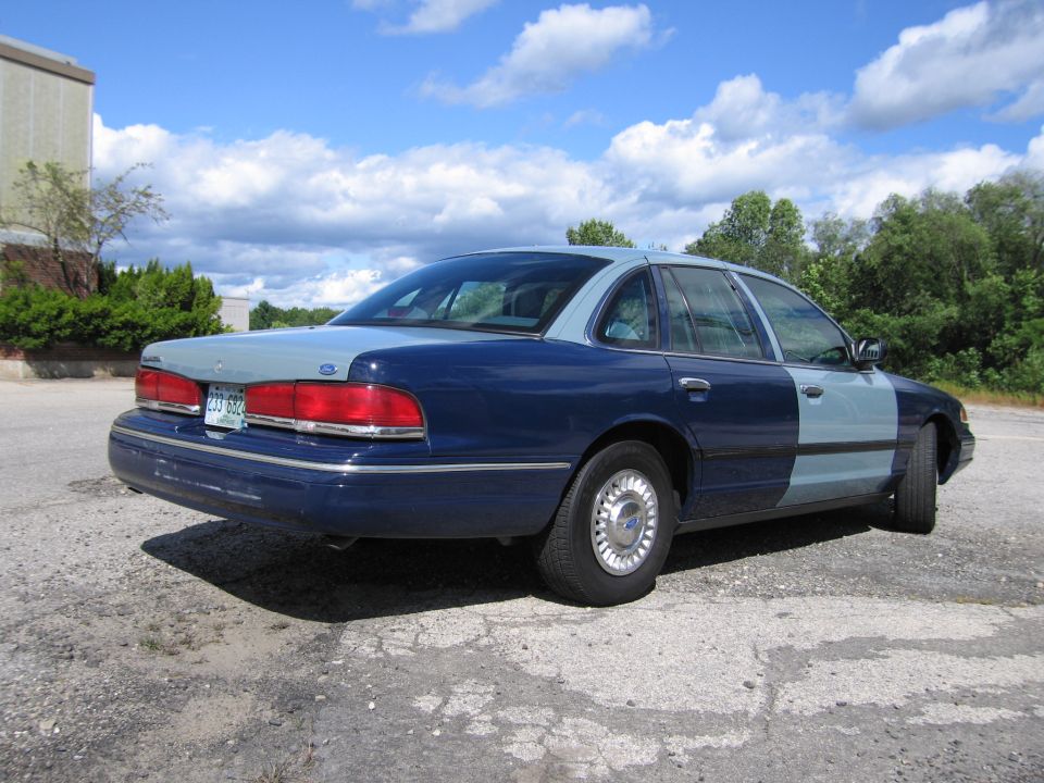 Two-tone blue Ford Crown Victoria sedan, rear quarter view on gravel lot