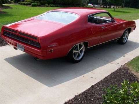 1978 Ford Bronco - A bright red 1970s era Ford Torino coupe is shown from a rear three-quarter angle, parked on a concrete driveway next to a grassy lawn.