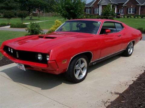 1978 Ford Bronco - A bright red 1970s Ford Torino GT coupe is parked on a concrete driveway in front of a brick house. The car features a black grille, hood s…