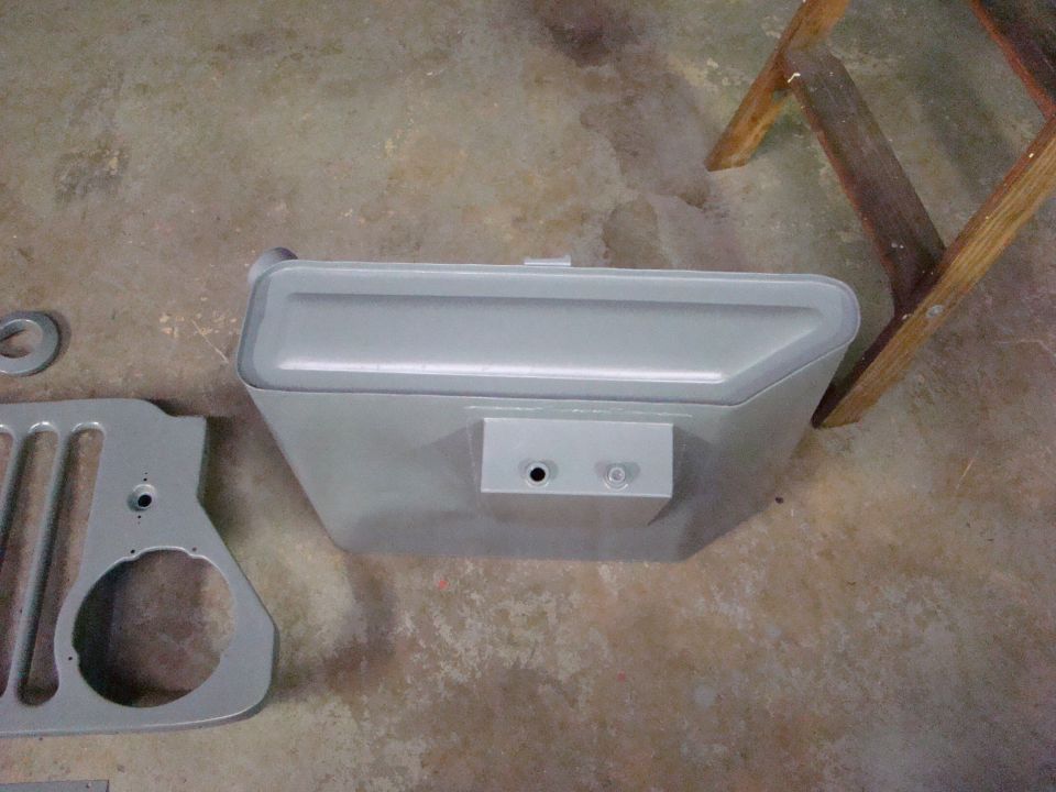 Gray painted Jeep fuel tank and grille section on concrete floor with wooden ladder.