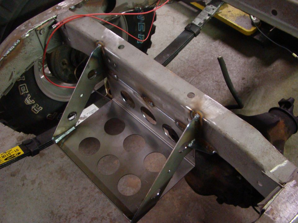 Custom welded metal battery tray attached to vehicle frame near tire and leaf spring.