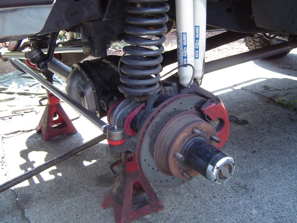 Front axle with wheel removed showing drilled rotor, red caliper, coil spring and dual white Rancho shocks