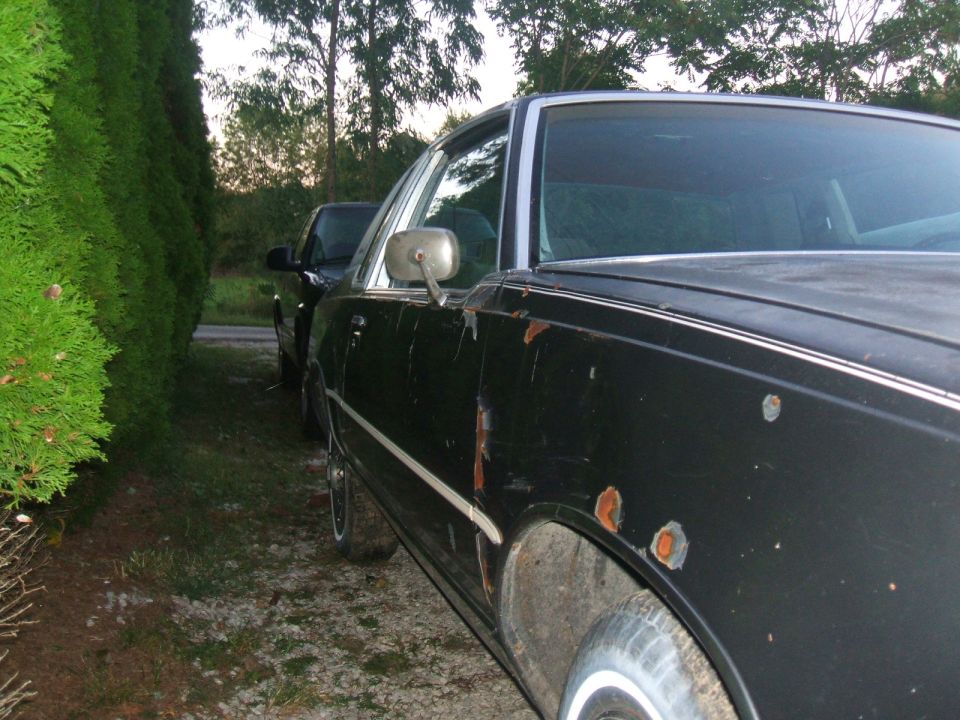 Black coupe with peeling paint and rust spots on rear quarter, parked on gravel by hedge