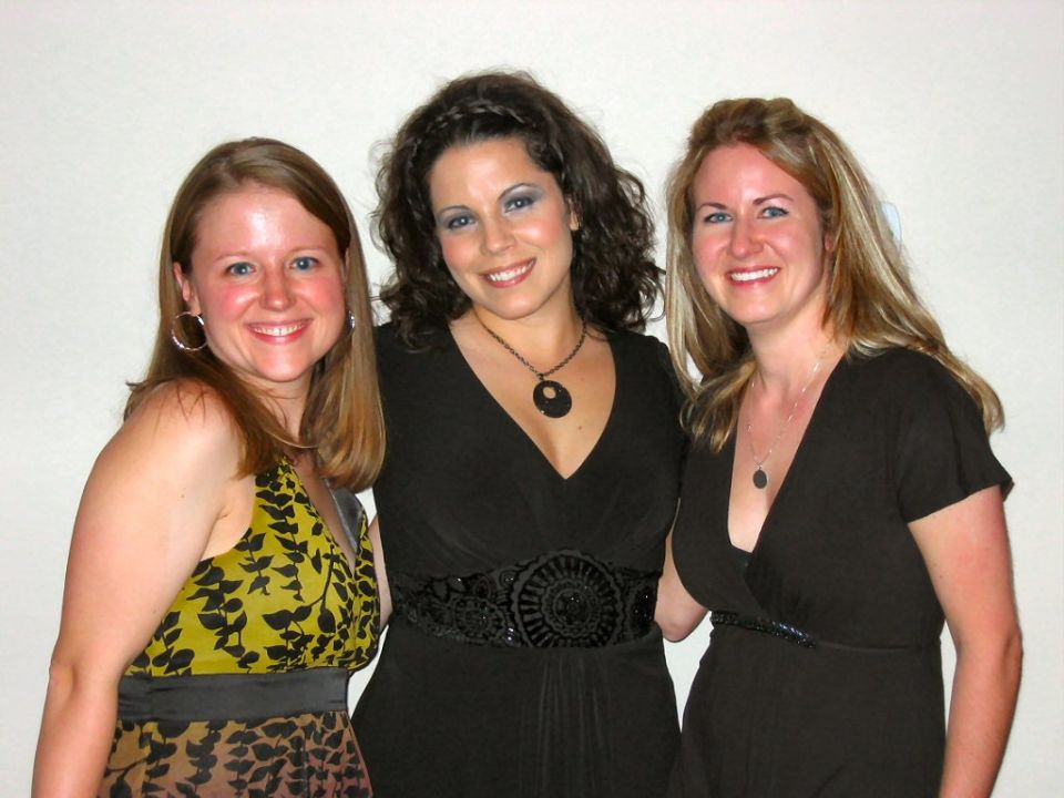 Three smiling women posing indoors against a plain wall