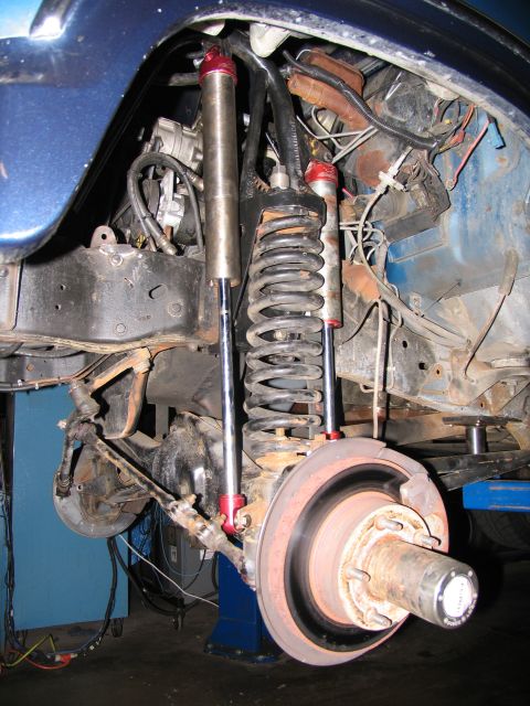 1984 Ford Bronco - The front suspension and brake assembly of a blue vehicle, likely a 1984 Ford Bronco, is shown with aftermarket shocks and large coil sprin…