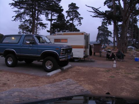 1984 Ford Bronco - A blue Ford Bronco is parked next to a white and orange travel trailer at a campsite. The Bronco is viewed from the front 3/4 angle, showin…