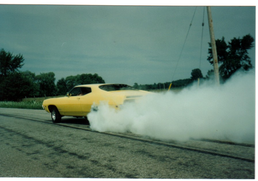 Yellow Ford Torino fastback doing a burnout with heavy white tire smoke on a rural road