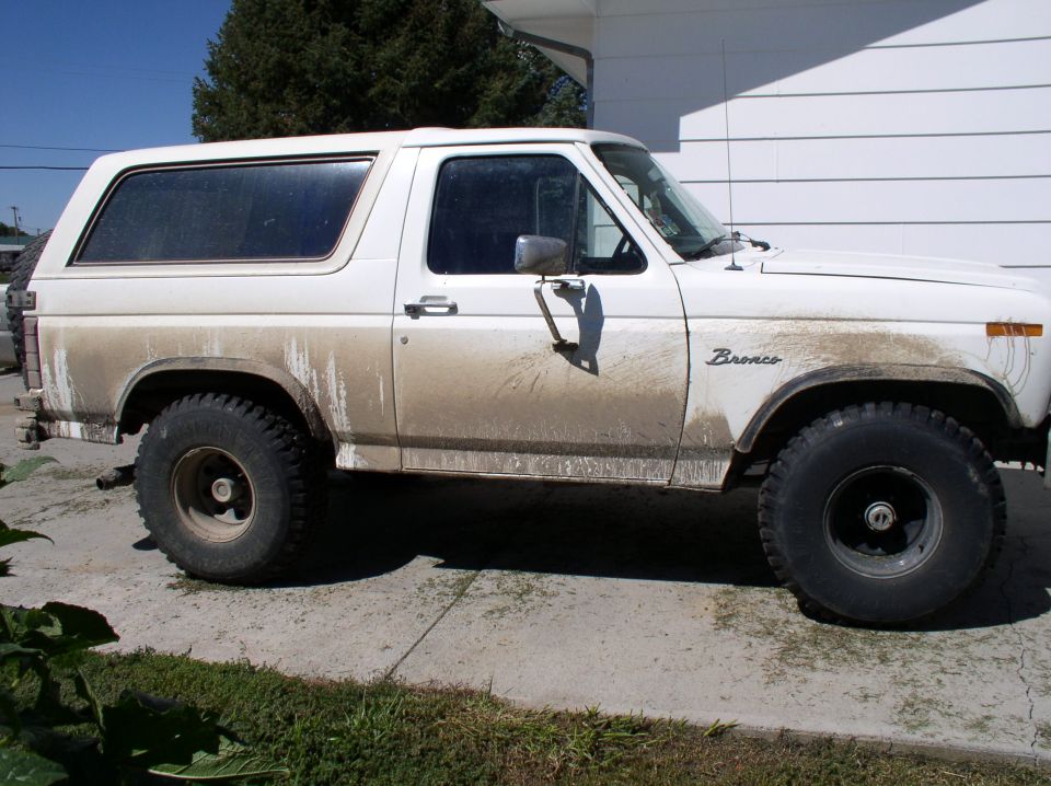 White two-door Ford Bronco SUV, side view, muddy lower body, parked on driveway.