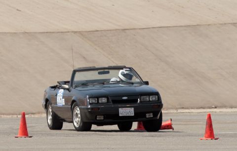 1985 Ford Mustang - A black 1985 Ford Mustang convertible with a driver wearing a white helmet navigates a course marked by orange traffic cones.