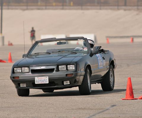 1985 Ford Mustang - A dark gray 1985 Ford Mustang convertible is shown in a three-quarter front view on a paved surface, possibly a race track, with orange con…