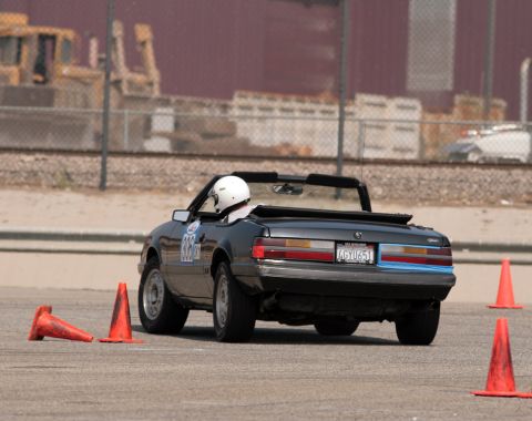 1985 Ford Mustang - A black convertible Ford Mustang is shown from the rear, driving on a paved surface with orange traffic cones. The driver is wearing a whit…