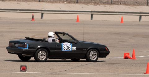 1985 Ford Mustang - A black convertible Mustang is participating in a timed event, indicated by the number 306 on its door and the orange cones on the asphalt…