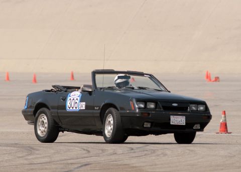 1985 Ford Mustang - A black 1985 Ford Mustang convertible is shown from the front 3/4 angle on a paved surface with orange cones visible in the background. The…