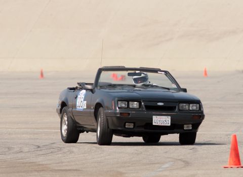 1985 Ford Mustang - A black 1985 Ford Mustang convertible is shown from the front at a race track, with a driver wearing a helmet visible inside. The car has a…