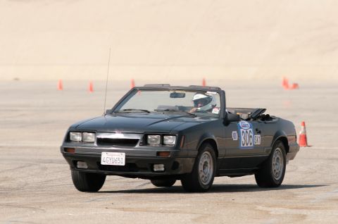 1985 Ford Mustang - Black Fox-body Ford Mustang GT convertible running an autocross course with the top down, driver in a white helmet, caught mid-turn between…