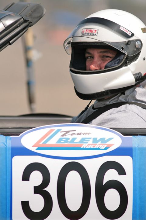 1985 Ford Mustang - A driver in a white G-Force Racing Gear full-face helmet with the visor up sits in an open-top car at an autocross, seen over the door. The…