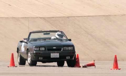 1985 Ford Mustang - A black convertible Ford Mustang is positioned on a paved surface, with the driver wearing a white helmet. Several orange traffic cones are…