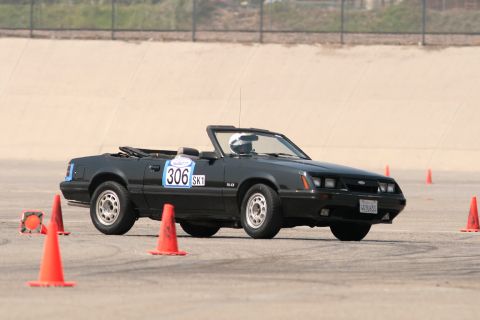 1985 Ford Mustang - Black Fox-body Mustang 5.0 convertible cornering hard through an autocross course, top down with a helmeted driver, roll bar and white '306…