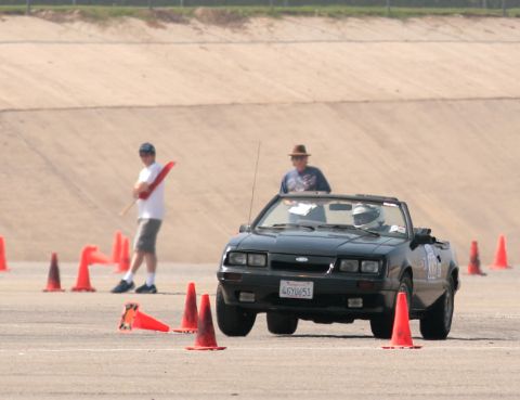 1985 Ford Mustang - A black convertible Mustang is shown on a paved track with orange cones. A person in a helmet is driving the car, and another person holdin…
