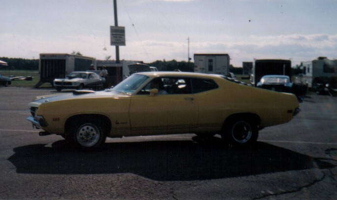 Yellow 1970 Ford Torino fastback in side profile with hood scoop, parked on pavement with trailers behind