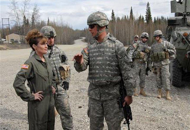 A woman in a green flight suit talks with soldiers in camouflage uniforms.