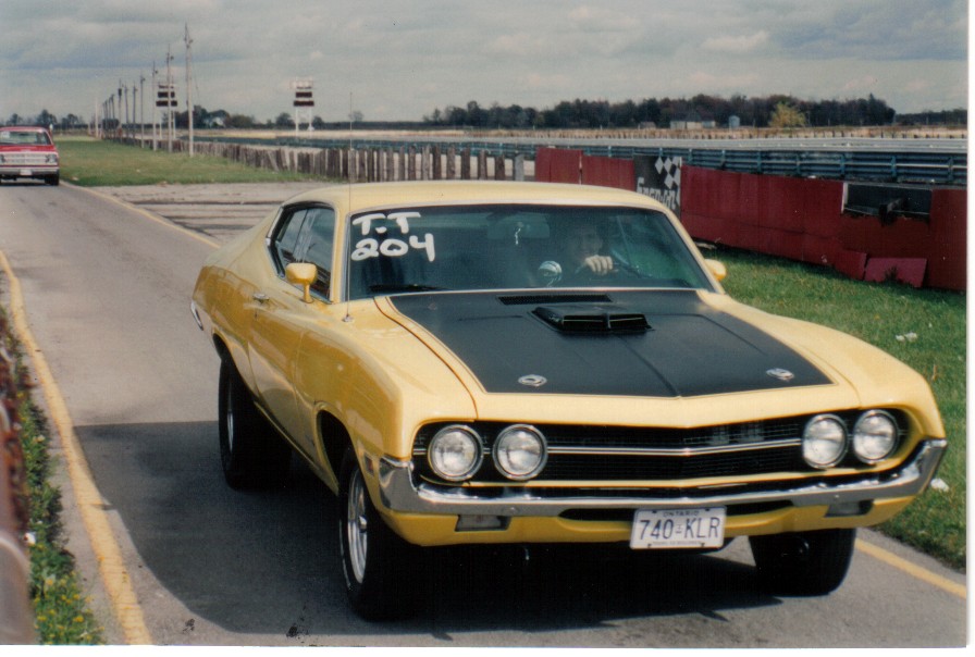Yellow 1970 Ford Torino with black hood and scoop, 'T.T 204' on windshield, at a drag strip