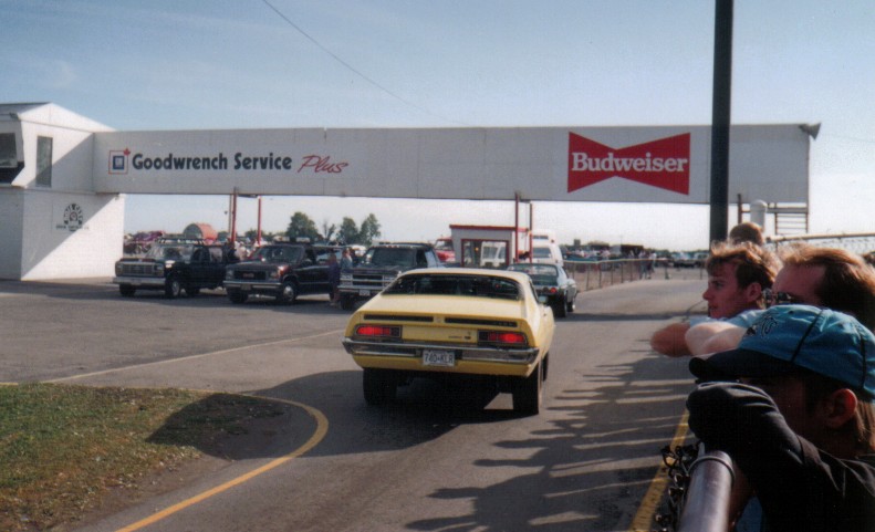 Yellow Ford Torino fastback from behind, driving under a Goodwrench/Budweiser bridge at a racetrack