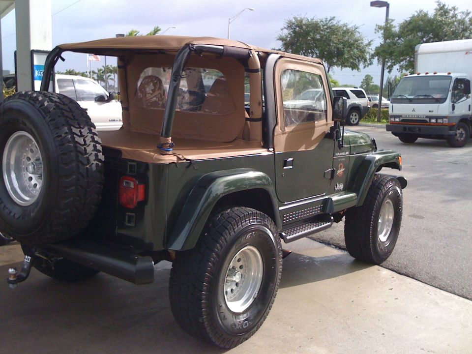 Dark green 1998 Jeep Wrangler with tan soft top, rear passenger side view, spare tire on back.