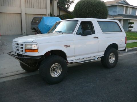 1988 Ford Bronco - A white 1988 Ford Bronco is parked on a residential street. The vehicle has a custom front bumper and large off-road tires.