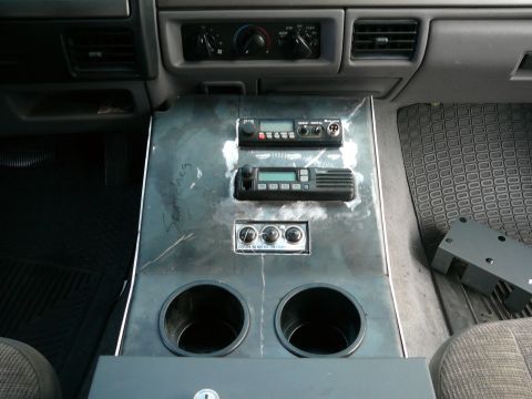 1988 Ford Bronco - The interior of a vehicle shows a custom metal console with two radios, three knobs, and two cup holders. A black rubber floor mat is visib…