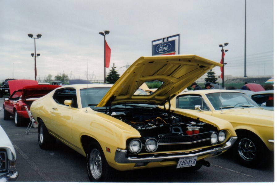 Yellow Ford Torino with hood open at a car show in front of a Ford dealership sign