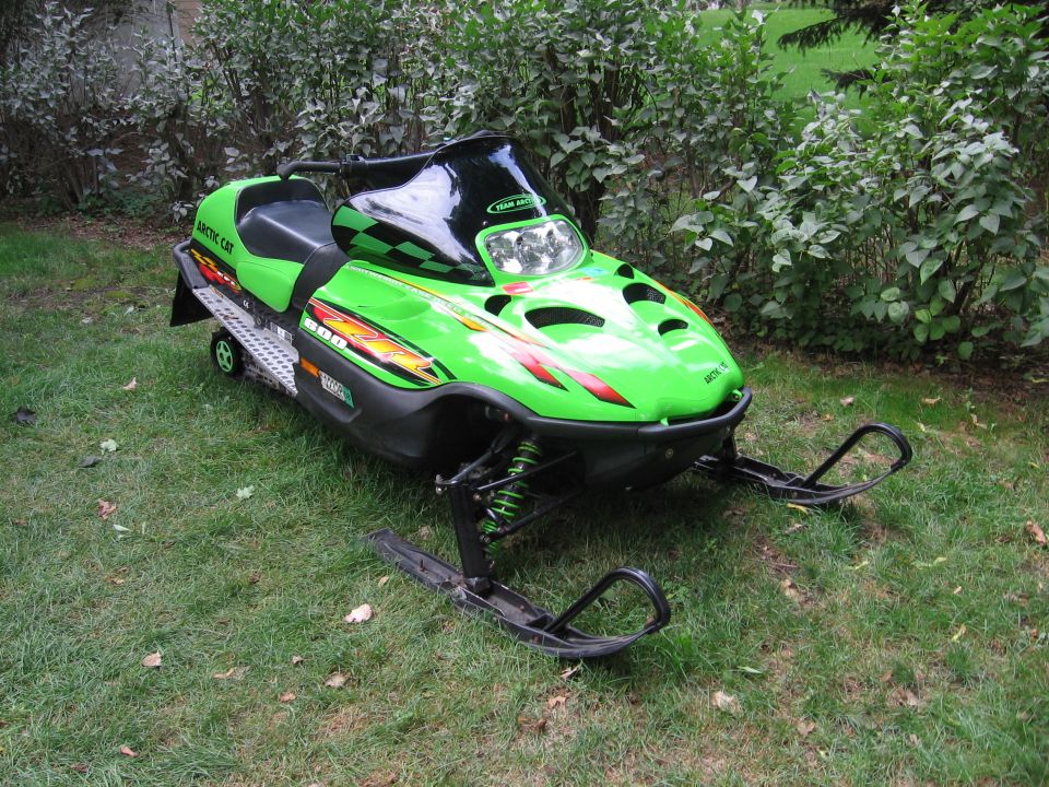 Green and black Arctic Cat snowmobile parked on grass beside shrubs